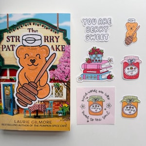 May include: A book titled "The Strawberry Patch Bake" with a bear-shaped sticker holding a honey dipper. Also, stickers with text "You are berry sweet", a jar of jam, and honey jars.