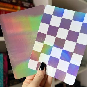 Könnte beinhalten: Zwei rechteckige holografische Aufkleber. Einer hat einen Regenbogen-Schimmer, der andere ein lila-weißes Schachbrettmuster. Die Aufkleber werden von einer Hand mit schwarzem Nagellack gehalten.