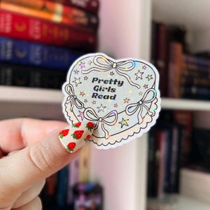May include: A holographic sticker shaped like a heart-shaped cake with the text "Pretty Girls Read" in black lettering. The sticker has a white background with a light blue frosting design and stars.