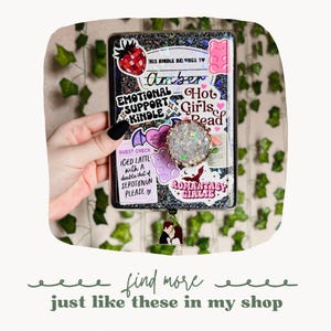 May include: A black and silver glitter covered notebook with a variety of colorful stickers, including a strawberry, a gummy bear, a bat, and a heart. The text "THIS KINDLE BELONGS TO Amber" is written on the cover. The notebook has a silver charm attached to it.