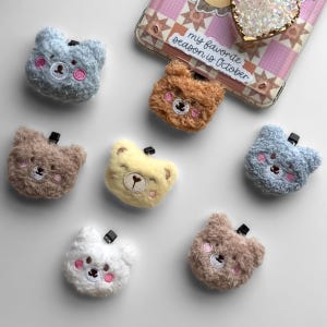 May include: Seven plush bear-shaped hair clips in various colors including light blue, brown, yellow, and white. Each clip features embroidered facial details with pink cheeks and a small black clip attachment. A phone case with the text "my favorite season is October" is visible in the corner.