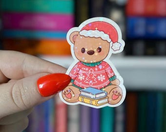 May include: A sticker featuring a brown teddy bear wearing a red Christmas sweater and Santa hat. The bear is sitting on top of two books. The sticker has a glittery, holographic effect.