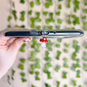 May include: A clear phone case with a red and white mushroom charm attached to the headphone jack.