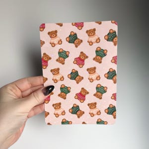 May include: A rectangular card with a repeating pattern of brown teddy bears wearing sweaters in various colors, including red, green, and cream, on a light pink background. The bears are arranged in a grid-like fashion, creating a playful and whimsical design.