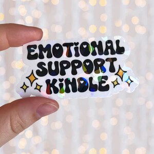 May include: A white iridescent sticker with black text that reads "Emotional Support Kindle" with gold glitter stars on either side.