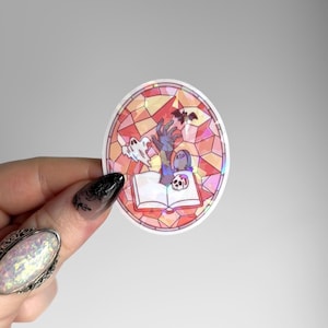 May include: An oval holographic sticker featuring a stained glass pattern in shades of pink and orange. The design depicts an open book with a hand reaching out, a small ghost, a bat, a skull, and a tombstone with the letters RIP inscribed.