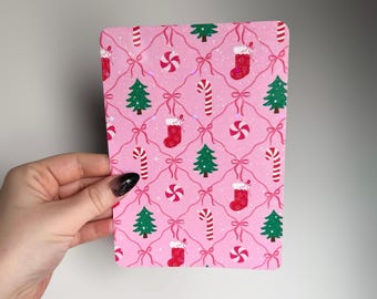 May include: A pink rectangular card with a repeating Christmas pattern. The pattern includes red stockings, green Christmas trees, candy canes, and pink bows. The card is held by a hand with a black and silver nail design.