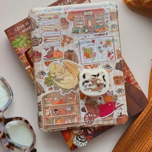 May include: A clear tablet case decorated with various autumn-themed stickers, including a sleeping yellow dragon, a red wheelbarrow, and a bookshelf. A white, cat-shaped phone grip with a clear, glitter-filled centre is attached to the case.
