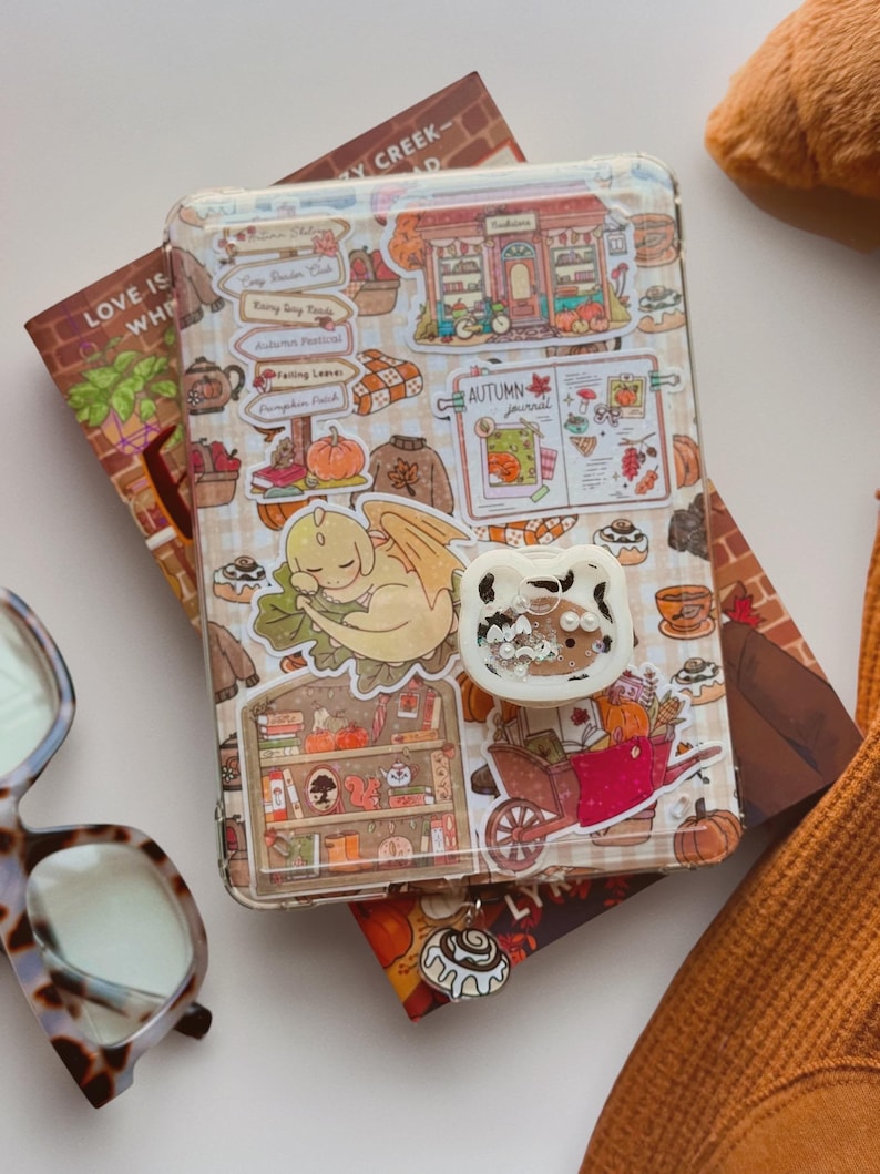 May include: A clear tablet case decorated with various autumn-themed stickers, including a sleeping yellow dragon, a red wheelbarrow, and a bookshelf. A white, cat-shaped phone grip with a clear, glitter-filled centre is attached to the case.
