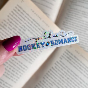 Puede incluir: Una pegatina blanca con acabado hologr&aacute;fico que muestra el texto "You had me at hockey romance" con un coraz&oacute;n y un disco de hockey.
