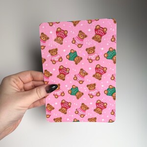 May include: A pink rectangular card with a repeating pattern of brown teddy bears wearing sweaters and hats in red, green, and pink. The background has a subtle star pattern.