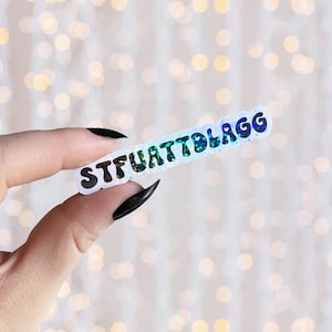 May include: A white sticker with the text "STFUATTBLAGG" in black, glittery letters. The sticker is iridescent and has a holographic effect.
