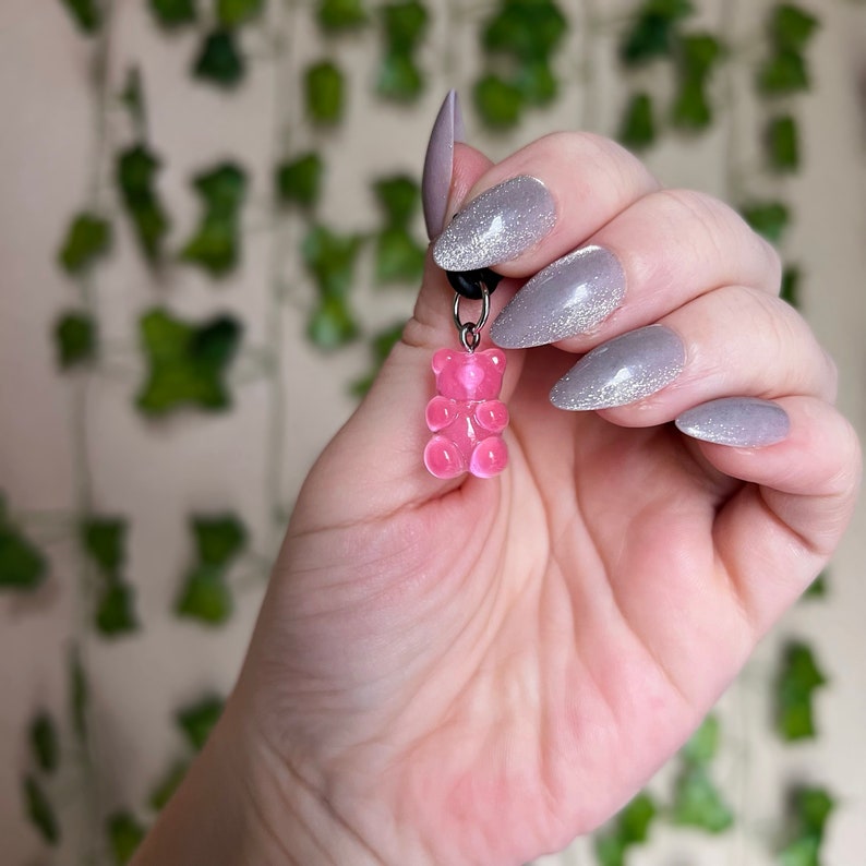 May include: A pink gummy bear charm on a silver chain. The charm is being held by a hand with gray nail polish.