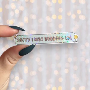 Op de afbeelding: Een holografische sticker met een lichtblauwe achtergrond en een witte rand. De sticker heeft een witte bloem en de tekst "sorry i was reading lol" in roze. Een gele smiley staat in de rechterbovenhoek.