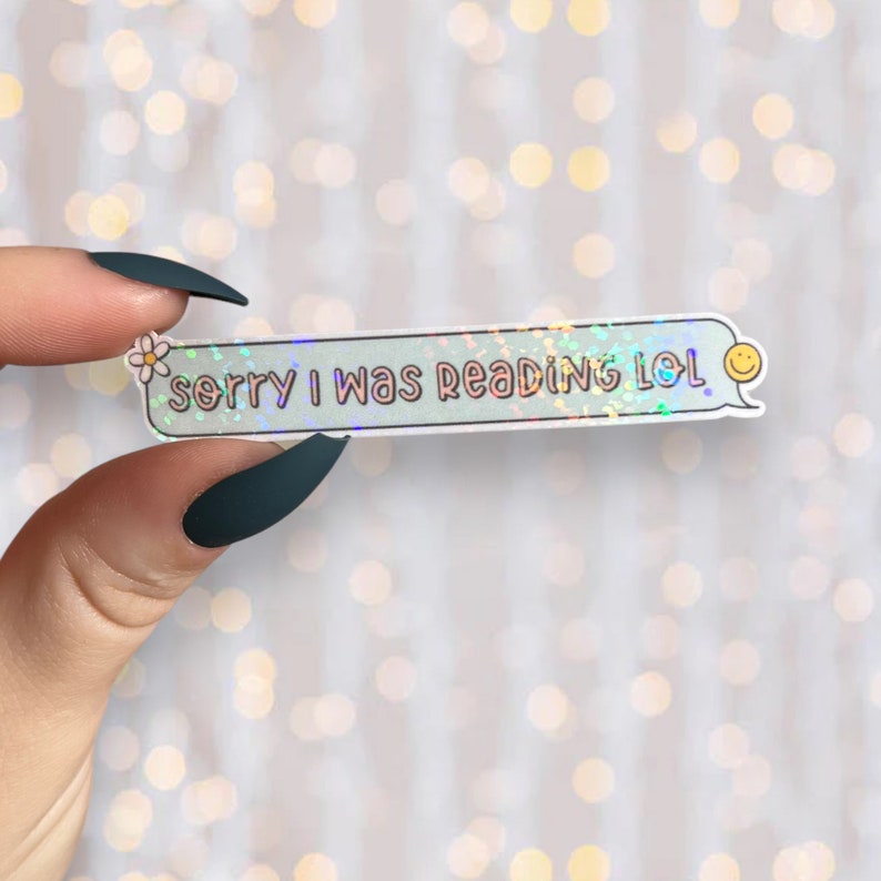Op de afbeelding: Een holografische sticker met een lichtblauwe achtergrond en een witte rand. De sticker heeft een witte bloem en de tekst "sorry i was reading lol" in roze. Een gele smiley staat in de rechterbovenhoek.