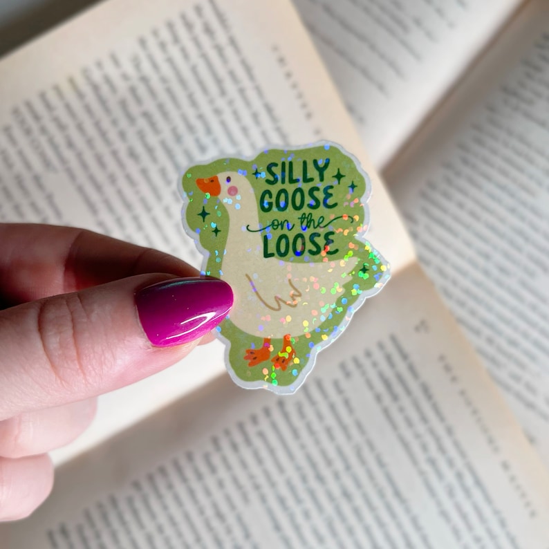 May include: A white goose with a green background and glitter. The goose has a red beak and feet. The text "Silly Goose on the Loose" is written on the sticker.