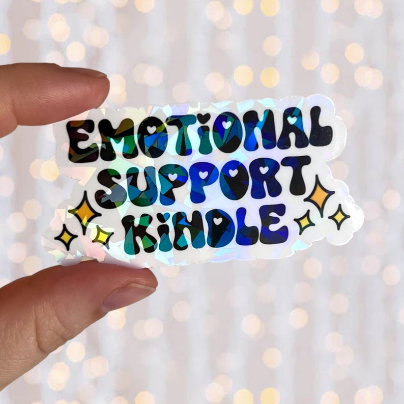 May include: A holographic sticker with the text "Emotional Support Kindle" in a rainbow font. The sticker has two yellow stars on either side of the text.