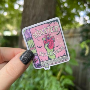 May include: A pink and green sticker with a book cover design. The book cover says "Monster Romance" and features a green hand holding a red heart.
