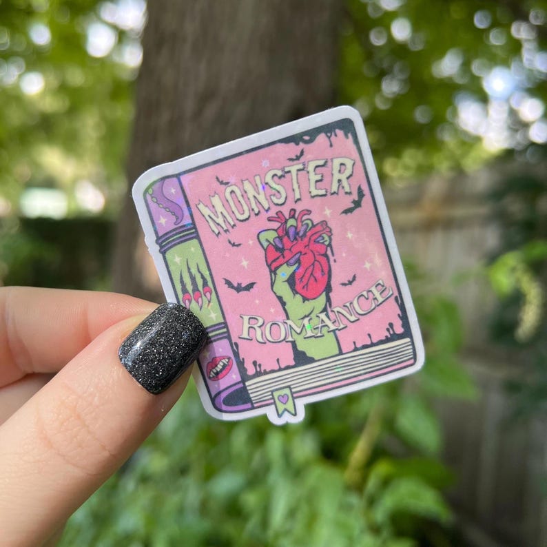 May include: A pink and green sticker with a book cover design. The book cover says "Monster Romance" and features a green hand holding a red heart.