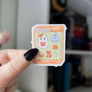May include: A sticker featuring a Cozy Ghoul theme. The sticker includes a ghost reading a book, a sweater with "Hello Fall", a pumpkin, a fox, a candle, and a coffee cup. The sticker is in a rectangular frame with the text "The Reader Fall Collection".