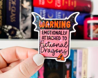 May include: A pink and black warning sticker with a glittery finish. The sticker reads "Warning: Emotionally attached to fictional dragons".