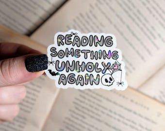 May include: A white sticker with the text "Reading something unholy again" in black letters with a glittery effect. The sticker also features a spider, a bat, and a skull.