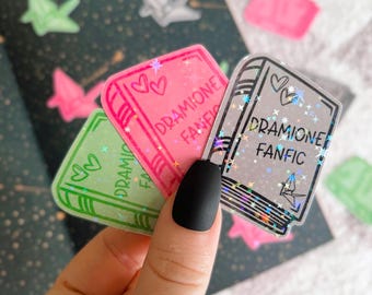 May include: Three clear holographic stickers with a book design. Each sticker features the text "DRAMIONE FANFIC" with a heart design. The stickers are on a white background.
