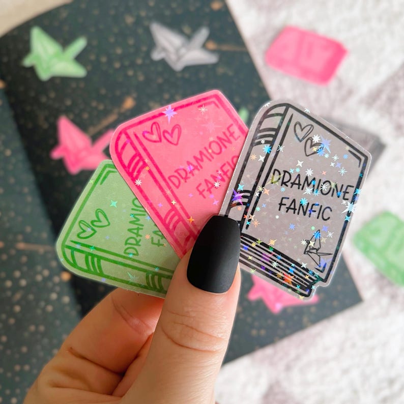 May include: Three clear holographic stickers with a book design. Each sticker features the text "DRAMIONE FANFIC" with a heart design. The stickers are on a white background.