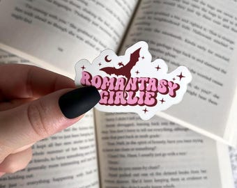 May include: A pink and red sticker with a white background featuring a black silhouette of a dragon with a crescent moon and stars. The sticker says "ROMANTASY GIRLIE" in a retro font.