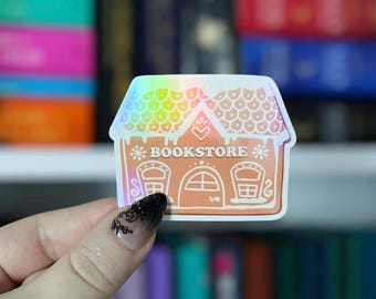 May include: A holographic sticker in the shape of a gingerbread house with the word "BOOKSTORE" printed on it. The sticker has a rainbow roof and a light brown body with two windows and a door.