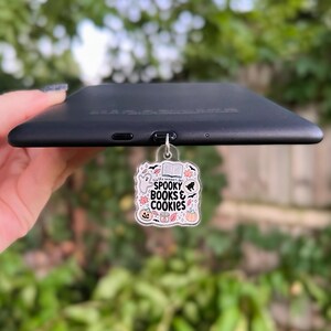 Puede incluir: Un charm de acrílico transparente con un diseño de libros y galletas espeluznantes. El charm presenta un fantasma, un libro, un gato, una calabaza y una taza de chocolate caliente. El charm está unido a un lector electrónico negro.