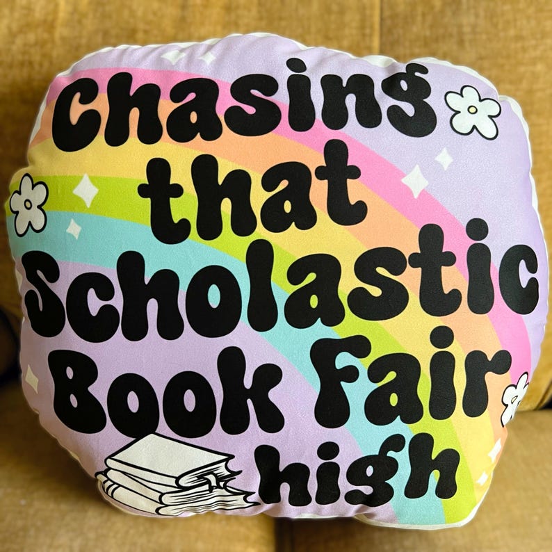 May include: A white pillow with a rainbow and stars design. The text "Chasing that Scholastic Book Fair high" is printed in black on the pillow.
