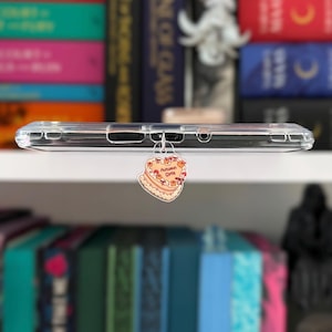 May include: A clear phone case with a heart-shaped charm. The charm features a cake design with the text "Autumn Cake" in a decorative font. The cake has orange and red accents, suggesting a fall theme.