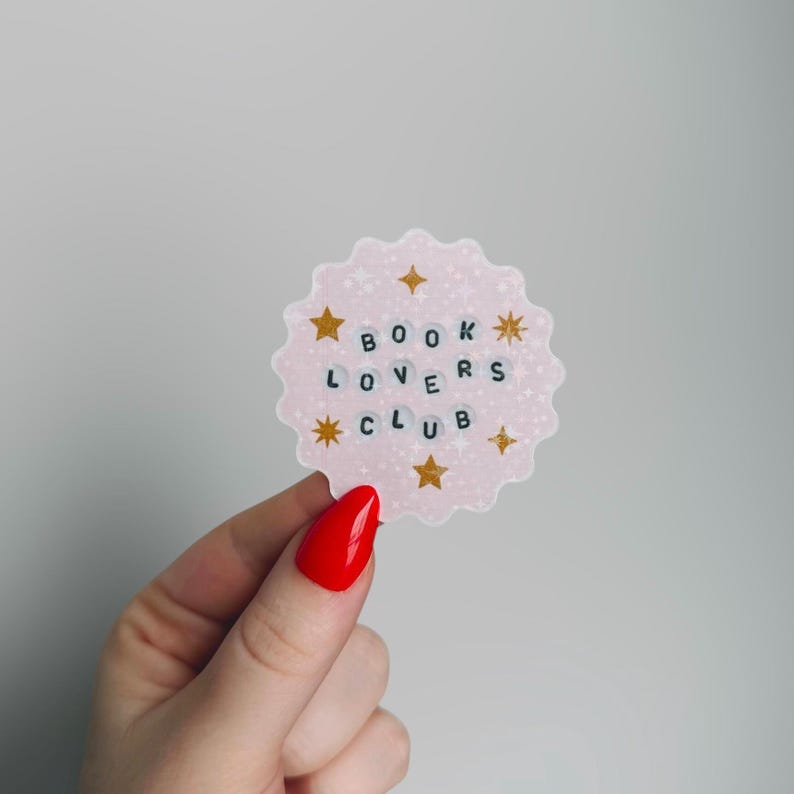 May include: A pink, scalloped sticker with the words "BOOK LOVERS CLUB" in black letter beads. Gold star and asterisk accents surround the text. The sticker is held by a hand with red nail polish.