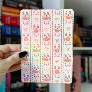Puede incluir: Una tarjeta rectangular con un patrón repetido de calabazas naranjas, flores rosas y lazos sobre un fondo crema. El diseño está dispuesto en rayas verticales con un borde festoneado. La tarjeta está sostenida por una mano con una uña pintada de negro.