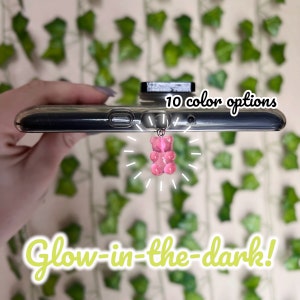Puede incluir: Un charm de teléfono con forma de osito de gominola rosa que brilla en la oscuridad, unido a una funda de teléfono transparente. 10 opciones de color.