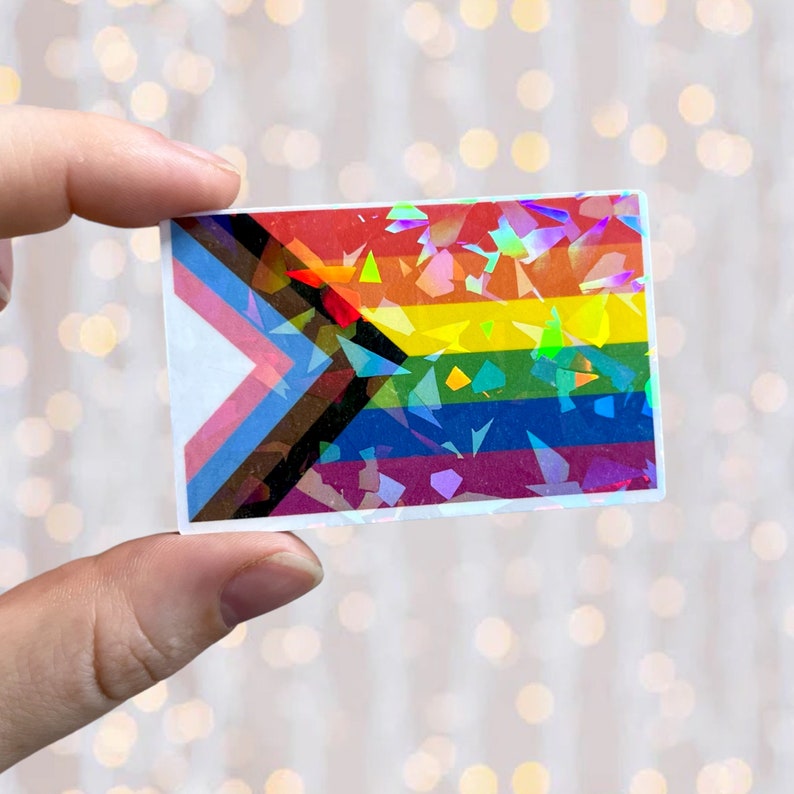 May include: A rainbow pride flag sticker with a black and brown triangle in the corner. The sticker has a holographic finish and is made of vinyl.
