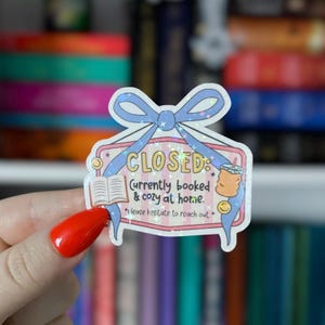 Puede incluir: Una pegatina decorativa con un lazo azul claro y la palabra "CLOSED". La pegatina incluye el texto "Currently booked & cozy at home" e ilustraciones de un libro y una bebida.