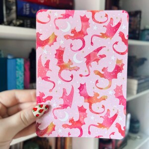 以下が含まれることがあります： ピンクのカードに、金色と白のアクセントがついたピンクのドラゴンシルエットの繰り返しパターンが描かれています。ドラゴンは飛んでおり、水彩画のような効果があります。