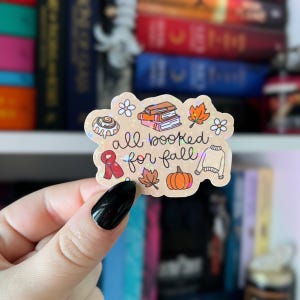 May include: A sticker with the text "all booked for fall" and fall-themed illustrations. The sticker features a cinnamon roll, books, leaves, a pumpkin, a scarf, a sweater, and flowers. The sticker has a holographic effect.