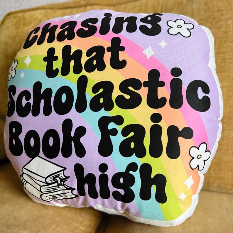 May include: A purple and pink pillow with a rainbow design and the text "chasing that scholastic book fair high" in black. The pillow has a stack of books on the bottom left corner.