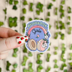 May include: A sticker with a blue background and a pink and green rainbow that says "Audiobook Girlie". The rainbow is over a pair of headphones with white flowers and a pink flower in the middle. The headphones are white and beige.