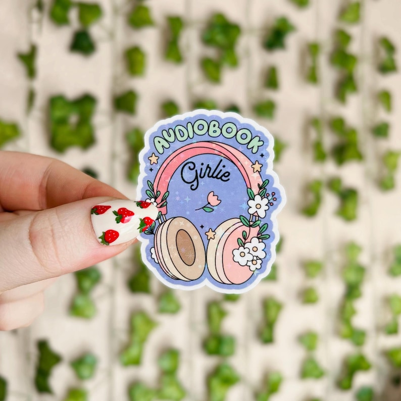 May include: A sticker with a blue background and a pink and green rainbow that says "Audiobook Girlie". The rainbow is over a pair of headphones with white flowers and a pink flower in the middle. The headphones are white and beige.