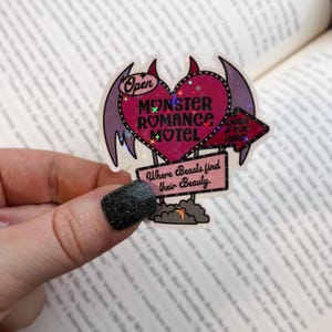May include: A pink heart-shaped sign with the text "Monster Romance Motel" and "Where Beasts Find Their Beauty". The sign has devil horns and wings and is decorated with glitter.