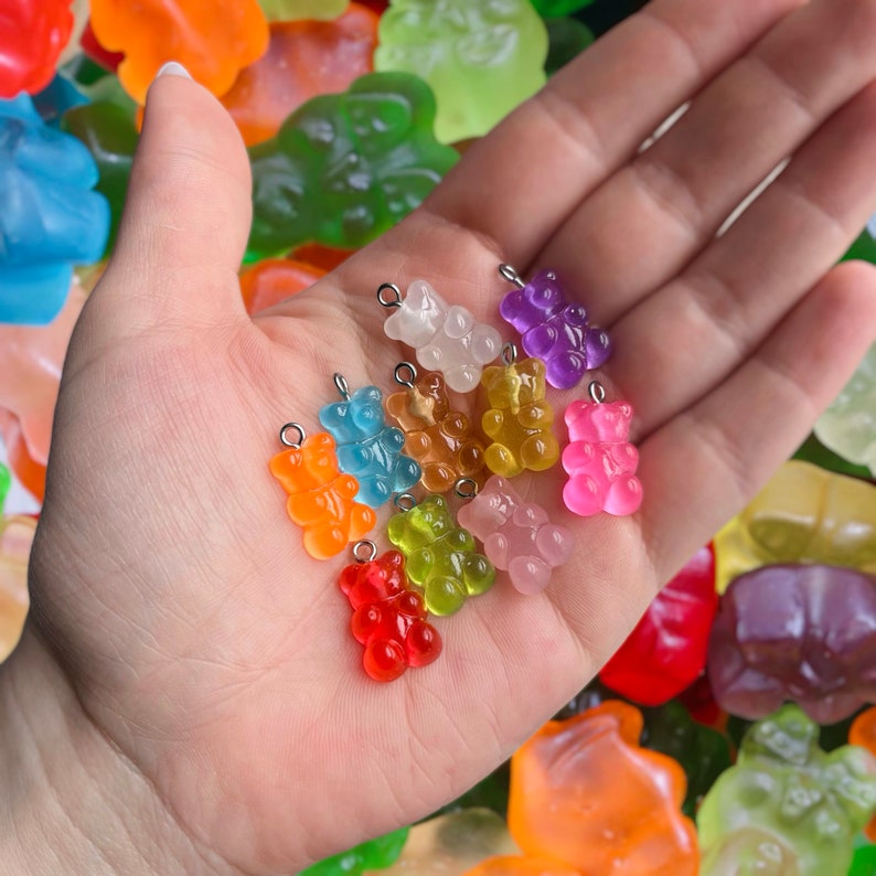May include: A collection of ten gummy bear charms in various colors, including red, orange, yellow, green, blue, purple, and pink. The charms are made of clear plastic and have a shiny finish. They are arranged on a hand, with the gummy bears facing up.