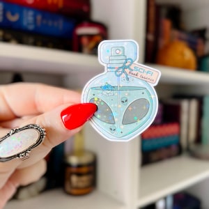 May include: A light blue sticker in the shape of a perfume bottle, labeled "SCI-FI Book Essence." The bottle contains a teal liquid with alien heads and a small spaceship. The sticker has a holographic, glittery effect.