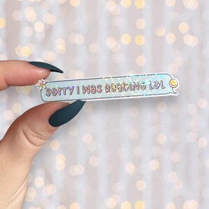 Op de afbeelding: Een holografische sticker met een blauwe achtergrond en witte tekst die "Sorry, ik was aan het lezen lol" zegt. Een kleine witte bloem en een gele smiley staan ​​aan weerszijden van de tekst.