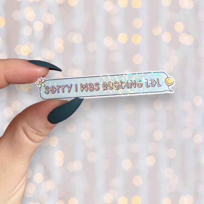 Op de afbeelding: Een holografische sticker met een blauwe achtergrond en witte tekst die "Sorry, ik was aan het lezen lol" zegt. Een kleine witte bloem en een gele smiley staan ​​aan weerszijden van de tekst.