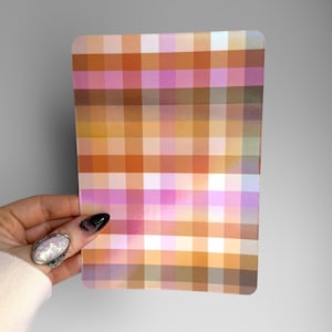 May include: A rectangular sheet featuring a repeating grid pattern of squares in various shades of orange, pink, white, and brown. The surface has a smooth, glossy finish that reflects light, creating a subtle iridescent sheen across the colours.