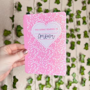 May include: Pink notebook-style cover with a white heart and the text "This Kindle belongs to Amber".